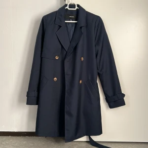Mörkblå trenchcoat från Vero Moda - Snygg mörkblå trenchcoat från Vero Moda med klassisk design. Jackan har långa ärmar och knappar framtill samt ett bälte i midjan för en elegant look. Perfekt för vår och höst.