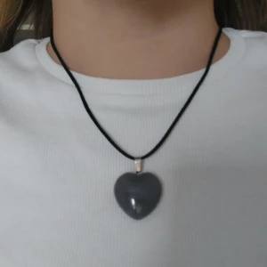 Halsband med hjärtformat hänge - Snyggt halsband med ett hjärtformat hänge i grå kristall. Hänget är fäst på en svart rem med ett enkelt lås.