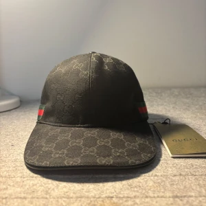 Svart Gucci keps - Snygg svart keps från Gucci med diskret mönster och klassiska gröna och röda ränder på sidan. Kepsen har en justerbar rem baktill för perfekt passform. Storlek ”M”
