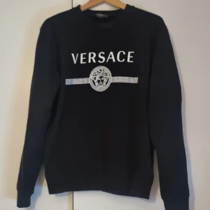 Svart tröja från Versace - Säljer en svart tröja från Versace med deras ikoniska logga och mönster framtill. Tröjan har långa ärmar och en klassisk rund halsringning. Perfekt för en stilren look. HELT ÄKTA. KÖPT FRÅN NK. Minimalt hål på axlen som kan ses på tredje bilden. 4800 orginal pris.