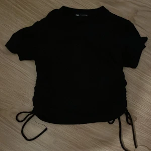 Svart kortärmad topp från Zara - Säljer en svart kortärmad topp från Zara med snörning i sidorna. Perfekt för en stilren look. Tillverkad i ett mjukt material som ger en bekväm passform. Storleken är mindre än vanligt