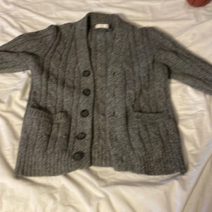 Grå stickad kofta från Bläck inga  skador  - Säljer en stilren grå stickad kofta från Bläck med knappar framtill. Koftan har ett klassiskt flätmönster och två praktiska fickor. Perfekt för kyliga dagar!
