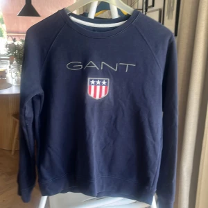 Mörkblå sweatshirt från GANT - Säljer en stilren mörkblå sweatshirt från GANT med deras ikoniska logga och emblem på bröstet. Tröjan har långa ärmar och en rund halsringning. Perfekt för en avslappnad och snygg look.