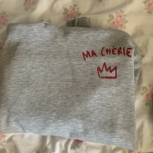 Grå tröja från Gina Tricot - Säljer en grå tröja från Gina Tricot med texten 'Ma Chérie' och en krona i rött tryck på bröstet. Perfekt för en avslappnad stil. Tröjan är mjuk och bekväm.