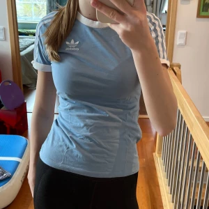 Ljusblå t-shirt från Adidas - Säljer en snygg ljusblå t-shirt från Adidas med klassiska vita ränder på ärmarna och vit krage. Perfekt för en sportig look eller vardagsstil. Adidas-loggan är tryckt på bröstet. Passar perfekt till jeans eller som träningströja!