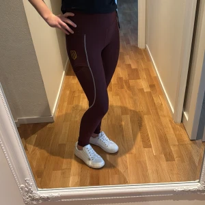 Vinröda ridbyxor från Maya Delorez - Snygga vinröda ridbyxor från Maya Delorez med hög midja och coola mönsterdetaljer längs benen. Perfekta för träning eller en aktiv dag. Tillverkade i en skön mix av polyester och spandex för bästa komfort och passform.