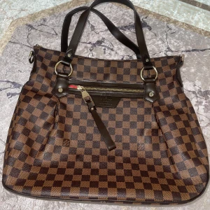 Brun rutig axelväska från Louis Vuitton - Snygg brun axelväska från Louis Vuitton med klassiskt rutigt mönster. Väskan har en praktisk dragkedja och eleganta detaljer i skinn. Perfekt för att bära dina dagliga nödvändigheter med stil.