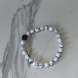 Vitt armband med marmorerade pärlor - Snyggt armband med vita marmorerade pärlor och en svart detalj. Perfekt för att ge en stilren touch till din outfit. Elastiskt band för enkel passform.☀️