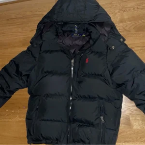 Svart dunjacka från Polo Ralph Lauren - Snygg svart dunjacka från Polo Ralph Lauren med huva och dragkedja. Jackan har en klassisk design med det ikoniska röda logotypen på bröstet. Perfekt för kyliga dagar.