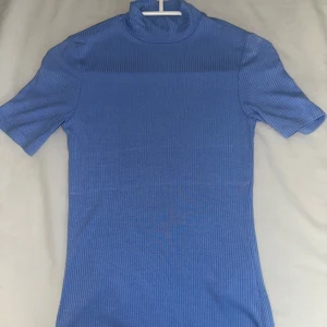 Blå ribbad polotröja - Säljer en snygg blå ribbad polotröja med korta ärmar. Perfekt för en stilren look. Tröjan har en hög krage och är gjord i ett mjukt material som sitter bekvämt.