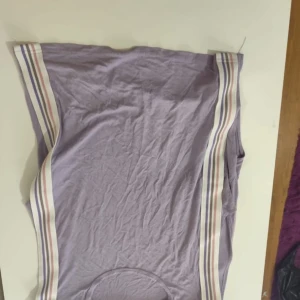 Lila ärmlös topp med randiga detaljer - Säljer en lila ärmlös topp med randiga detaljer i vitt, rosa och beige längs sidorna. Perfekt för en avslappnad stil.