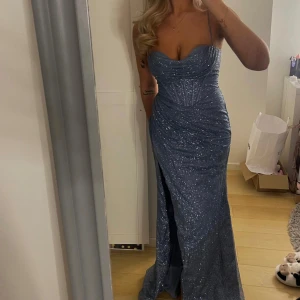 Blå glittrig långklänning - Säljer en fantastisk blå glittrig långklänning med smala axelband och snörning i ryggen. Klänningen har en elegant slits framtill och en figurnära passform som framhäver silhuetten. Köpt på promsandweddings i Malmö.