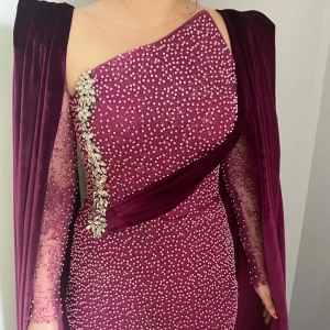 Vinröd glittrig långklänning - Elegant vinröd långklänning med glittrande detaljer och en axelbandslös design. Klänningen har långa ärmar och en vacker snörning i ryggen. Perfekt för en glamorös kväll. Underarmen på en sida har gått i sönder syns inte om man har axlarna på. Längden på klänningen är 155 cm. 