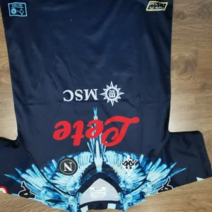 Mörkblå fotbollströja från Kappa - Snygg mörkblå fotbollströja från Kappa med coolt mönster i ljusblått och vita detaljer. Tröjan har korta ärmar och flera logotyper på framsidan. Perfekt för fotbollsfans som vill sticka ut på läktaren eller planen.