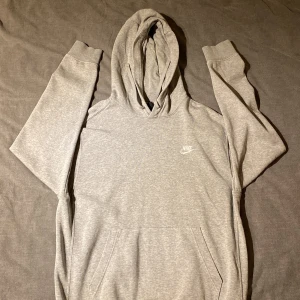 Grå hoodie från Nike - Säljer en klassisk grå hoodie från Nike med en liten logga på bröstet. Tröjan har en stor ficka framtill och en justerbar huva. Kan gå ner i pris vid snabb affär!