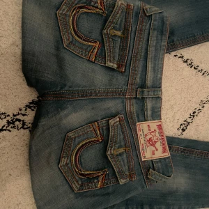 Blå jeans från True Religion - Säljer ett par snygga blå jeans från True Religion med coola detaljer på bakfickorna och kontrastsömmar. Jeansen har en klassisk femficksdesign och knappgylf. Perfekta för en avslappnad stil. Gylfen är dock trasig!!!