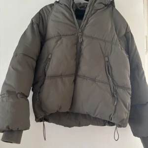 Grå/gröb pufferjacka med dragkedja - Säljer en stilren grå/grön pufferjacka med dragkedja och huva. Jackan har långa ärmar och praktiska fickor med dragkedjor. Perfekt för kyliga dagar.