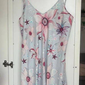 Blommig långklänning med smala axelband - Säljer en vacker långklänning med blommönster i rosa och lila nyanser på en ljusblå bakgrund. Klänningen har smala axelband och en v-ringad halslinje, perfekt för sommarens varma dagar.