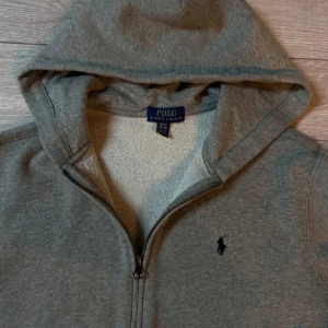 Ralph Lauren Hoodie - Säljer en grå hoodie från Ralph Lauren med dragkedja och huva. Den har en liten broderad logotyp på bröstet och är tillverkad i ett mjukt material som känns skönt mot huden. Perfekt för kyliga dagar. Sjukt bra skick. XL i barn sitter som S.