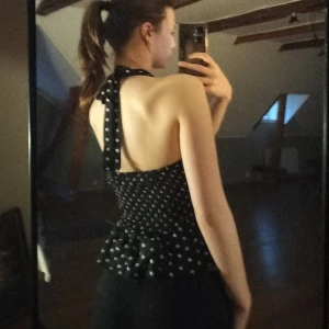 Svart prickig halterneck topp - Säljer en svart halterneck topp med vita prickar och volang nertill. Toppen har djup v-ringning och knyts i nacken. Perfekt för sommaren och ger en snygg retrostil. Aldrig använd bara testad. Klippte dock bort lappen då den kliade 