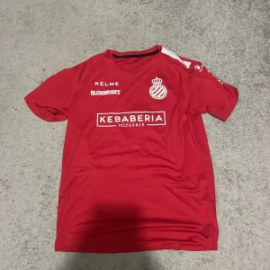 Röd RCD Espanyol fotbollströja från Kelme - Säljer en röd fotbollströja från Kelme med RCD Espanyol de Barcelona-logga på bröstet. Tröjan har vita tryck med sponsorloggor och klubbmärke, samt rund hals. Perfekt för dig som gillar spansk fotboll och vill sticka ut på planen.