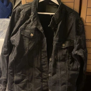 Svart jeansjacka med knappar - Säljer en svart jeansjacka med klassisk design och knappar framtill. Jackan har två bröstfickor med lock och knapp, samt markerade sömmar för en snygg look. Perfekt för dig som vill ha en tidlös och stilren jacka.