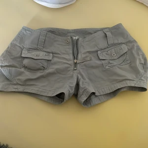 Ljusgrå/gröna shorts från J.Lindeberg - Snygga ljusgrå/gröna shorts från J.Lindeberg med två fickor framtill med lock och knapp, samt dragkedja och knappstängning. Perfekta för sommardagar och har en klassisk, stilren look.
