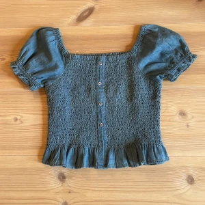 Blå smockad blus med puffärm från KappAhl - Supersöt blå jeans blus från KappAhl med smockad kropp, puffiga korta ärmar och volang nertill. Blusen har knappar framtill. Skön och luftig, passar till många outfits!