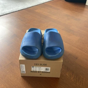 Blåa adidas Yeezy Slide sandaler - Säljer ett par blåa adidas Yeezy Slide sandaler. De har en chunky design med öppen tå och platt sula, tillverkade i syntetmaterial. Perfekta för sommaren och riktigt sköna att glida runt i. Boxen medföljer. Det är använd i ett halv år typ så lite slitna, original pris 1200kr. Tofflorna är lite små i storlek,fråga om ni undrar något 