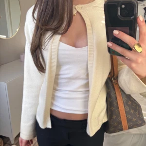 Vit kofta med knappdetalj - Säljer en stilren vit kofta med en enkel knapp upptill. Koftan är i mjukt material och har lång ärm. Perfekt att slänga över en topp för en clean look.