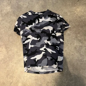 Camouflage t-shirt från Valentino - Säljer en snygg t-shirt från Valentino med grått, svart och vitt camouflage-mönster. T-shirten har korta ärmar och en liten metallnit bak vid nacken. Perfekt för dig som gillar streetstyle.