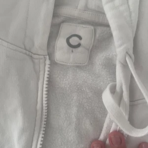 Vit hoodie för damer - Vit hoodie med dragkedja framtill och huva med snörning. Tröjan har två fickor framtill och är i mjukt material, perfekt för en avslappnad stil.