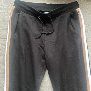 Svarta mjukisbyxor från BOSS med revärer - Svarta mjukisbyxor från BOSS med bruna och vita revärer längs sidorna. Byxorna har snörning i midjan och fickor fram samt en bakficka. Perfekta för en avslappnad stil. Dom är helt oanvända så inga skador eller märken på byxorna