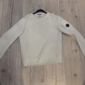 Vit stickad tröja från C.P. Company - Säljer en vit stickad tröja från C.P. Company med rund hals och ribbade muddar. Tröjan har en ikonisk svart linsdetalj på ena ärmen. Perfekt för dig som gillar stilrena och exklusiva plagg. Säljer denna vara billigare pga av att märket har börjat lossna plus ett litet hål där bak som knappt syns, kom gärna med bud!! Hör av er för frågor