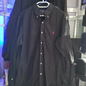 Ralph Lauren skjorta  - Säljer en svart skjorta från Ralph Lauren med klassisk krage och knappar framtill. Slim fit-modell i 100% bomull med stretch. Ikonisk röd broderad logga på bröstet. Perfekt för en stilren look. Den är xl i barnstorlek men skulle säga att den sitter som en L inge skador på den