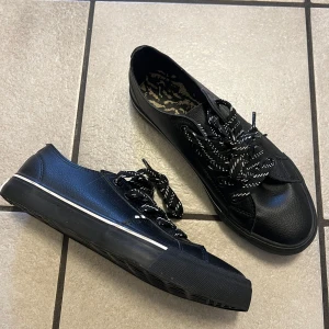 Svarta sneakers med mönstrade skosnören - Svarta sneakers med vit rand på sulan och svartvita mönstrade skosnören. Skorna har rund tå och platt sula. Insidan har ett djurmönstrat tyg. Perfekta för en avslappnad och stilren look.