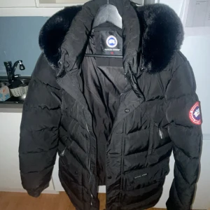 Svart dunjacka från Canada Goose med pälskrage - Säljer en svart dunjacka från Canada Goose med stor huva och pälskant. Jackan har dragkedja, knappar och flera fickor samt den klassiska loggan på ärmen. Perfekt för kalla vinterdagar.