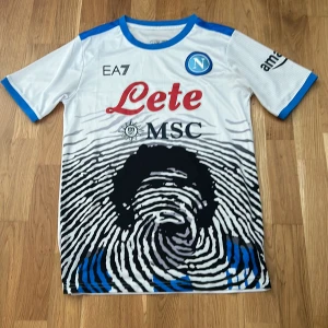 Napoli fotbollströja  - Säljer en vit Napoli fotbollströja från EA7 med maradona på magen, en riktigt fet fotbollströja som är perfekt nu till sommaren den är i bra skick!