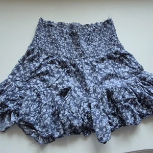 Blå blommig volangkjol - Supersöt blå kjol med blommigt mönster och volanger. Kjolen har bred resår i midjan för en bekväm passform och är perfekt för dig som gillar en feminin stil. Passar till både sneakers och sandaler.