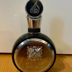 Lattafa Perfumes - Fakhar Black EdP - Parfym från Lattafa med elegant svart och silver flaska, dekorerad med ett detaljerat emblem och arabiska bokstäver. Doften har aromatiska, fräscht kryddiga, träiga och fruktiga noter samt inslag av citrus, lavendel och amber. Den är bara provsprejad.