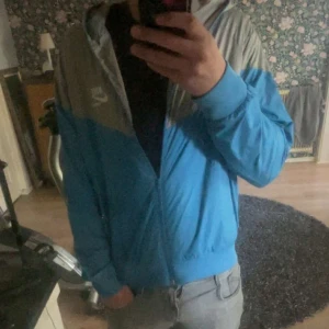 Blå och grå vindjacka från Nike - Säljer en snygg blå och grå vindjacka från Kappa med dragkedja framtill och huva. Jackan har långa ärmar och en sportig look, perfekt för utomhusaktiviteter. Kappas logga syns på bröstet.