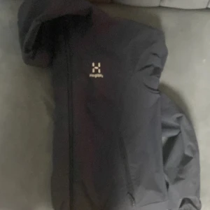 Svart vindjacka från Haglöfs - Säljer en svart vindjacka från Haglöfs med huva och dragkedja framtill. Jackan har en stilren design och är perfekt för blåsiga dagar. Klassisk logga på bröstet.