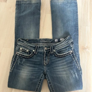 Bootcut jeans från miss me - Snygga blå jeans från Miss Me med broderade detaljer och dekorativa fickor både fram och bak. Byxorna har en klassisk bootcut-modell och tydliga slitningar för en cool look. Stängs med dragkedja och knapp. Midjemått 26.