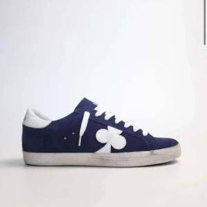 Blå sneakers från The Gallerist med vit klöverdetalj - Säljer ett par blå sneakers från The Gallerist, slutsålda! med vit klöverdetalj på sidan och vit häl med logotyp. Skorna har vita snören och sula, samt blå och vit mönstrad detalj bak. Köpta för 2400kr. De är lite slitna därav pris!