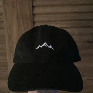 Svart keps med broderat bergmotiv - Svart keps med ett stilrent vitt broderat bergmotiv framtill. Klassisk böjd skärm och justerbar passform baktill.