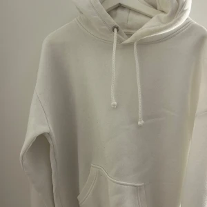 Vit hoodie med ficka - Säljer en stilren vit hoodie med huva och dragsko. Tröjan har en stor magficka och är perfekt för en avslappnad look. Passar både till jeans och mjukisbyxor.