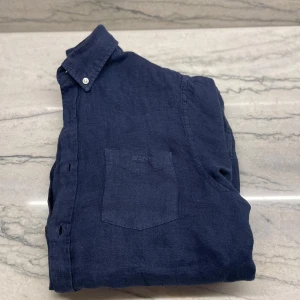 Mörkblå linneskjorta från Gant - Säljer en stilren mörkblå skjorta i linne från Gant. Skjortan har lång ärm, klassisk krage och bröstficka. Perfekt för dig som gillar en avslappnad men snygg look. Passar dig som är 12-14