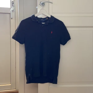 Svart pikétröja från Polo Ralph Lauren - Klassisk svart pikétröja från Polo Ralph Lauren med liten röd broderad logga på bröstet. Tröjan har krage, korta ärmar och knappar vid halsen. Perfekt för en stilren och avslappnad look.