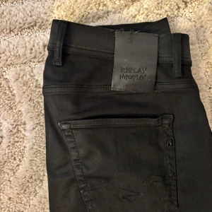 Svarta Replay Hyperflex jeans - Säljer ett par svarta Replay Hyperflex jeans med klassisk femficksdesign och stretchigt material. Byxorna har normal passform och raka ben, perfekt för en stilren look. Diskret Replay-logga på bakfickan och läderpatch i midjan. W30 L32 knappt andvända 