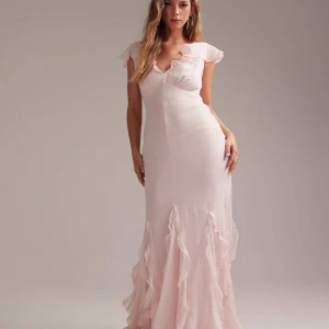 Ljusrosa långklänning med volangdetaljer - Romantisk ljusrosa långklänning med volangdetaljer vid axlar och nederkant. Klänningen har v-ringning, öppen rygg och är tillverkad i ett lätt, flytande material som ger en elegant siluett.